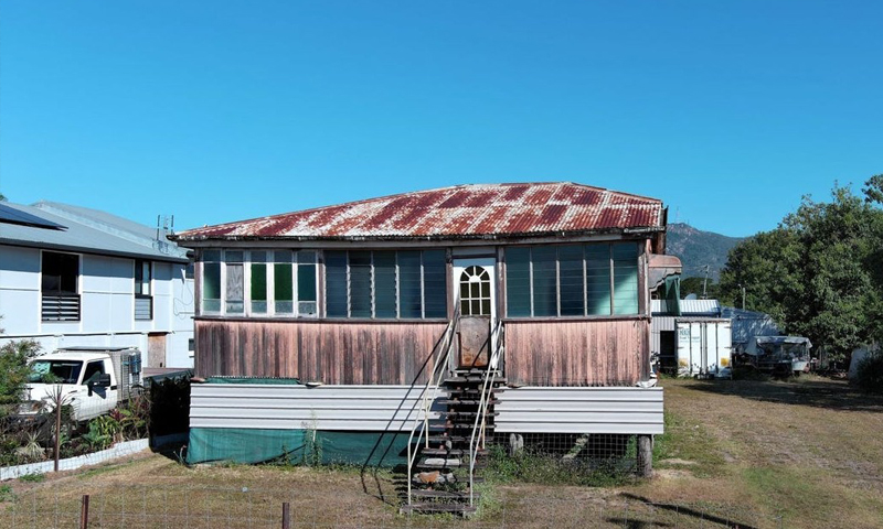 Old wooden house on stilts with a rusted red metal roof and a metal stairway leading to a front door, under a clear blue sky.