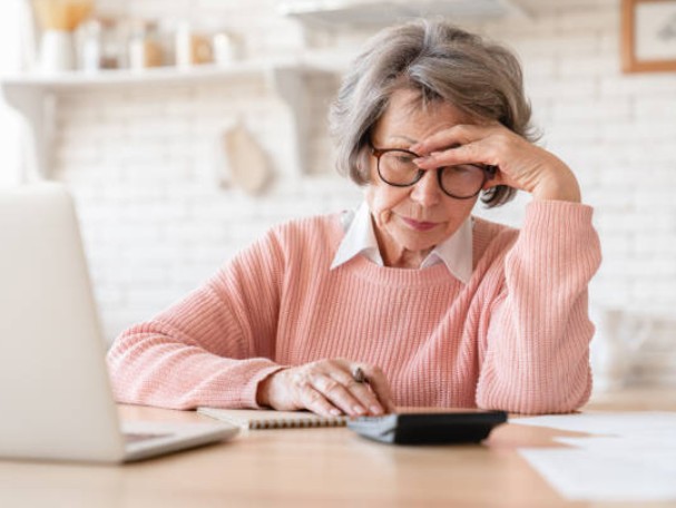 Older woman with gray hair and glasses sits at a desk, hand on her forehead, using a calculator and laptop in a bright home office, looking stressed.