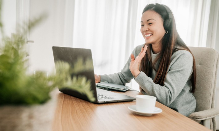 Woman with wireless headphones waving at her laptop during a video call at a wooden desk with a coffee cup nearby